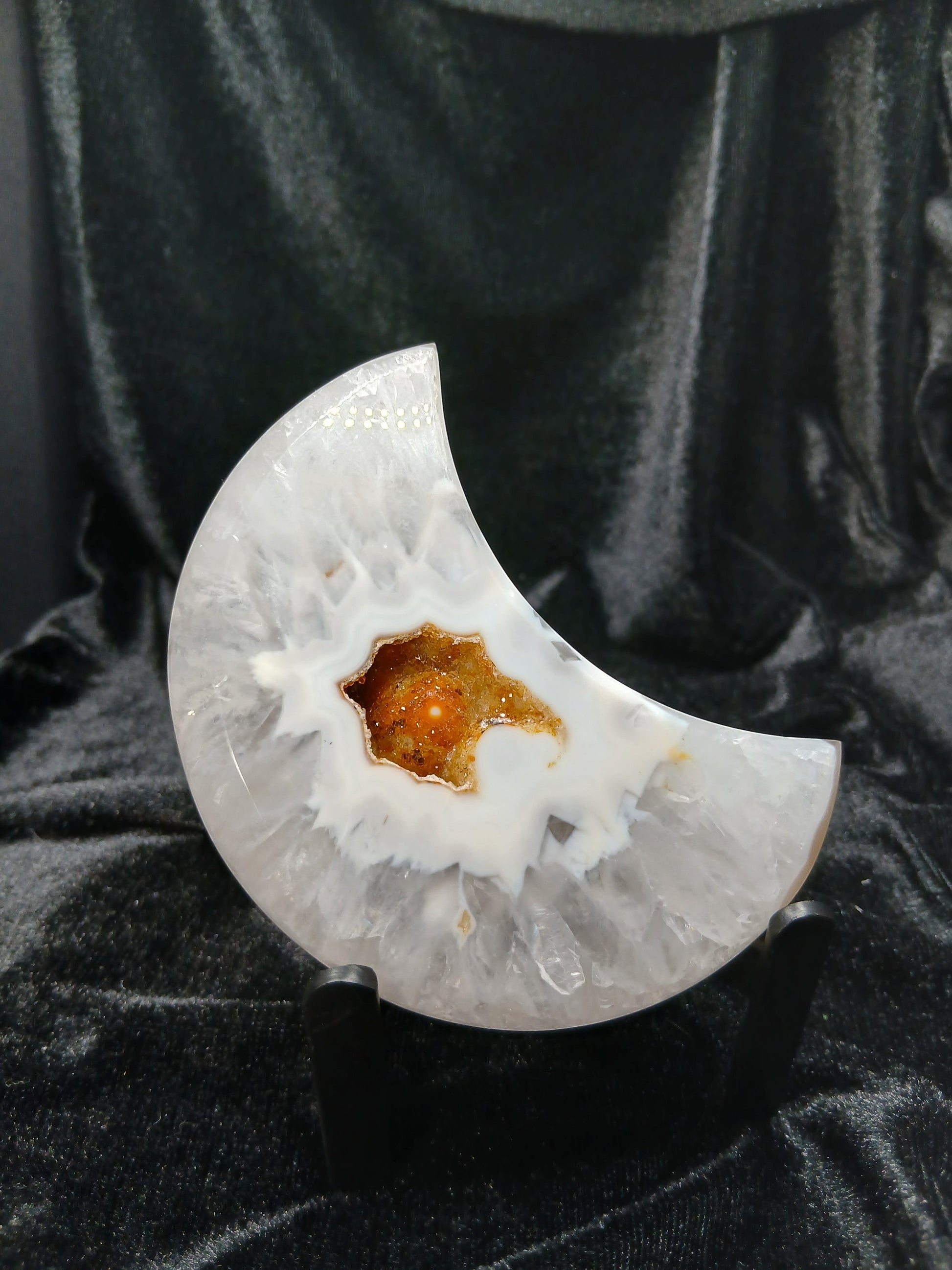 Fordite Haus: Crystals & Fordite agate moon carving with a central amber geode druzy on a black display stand.