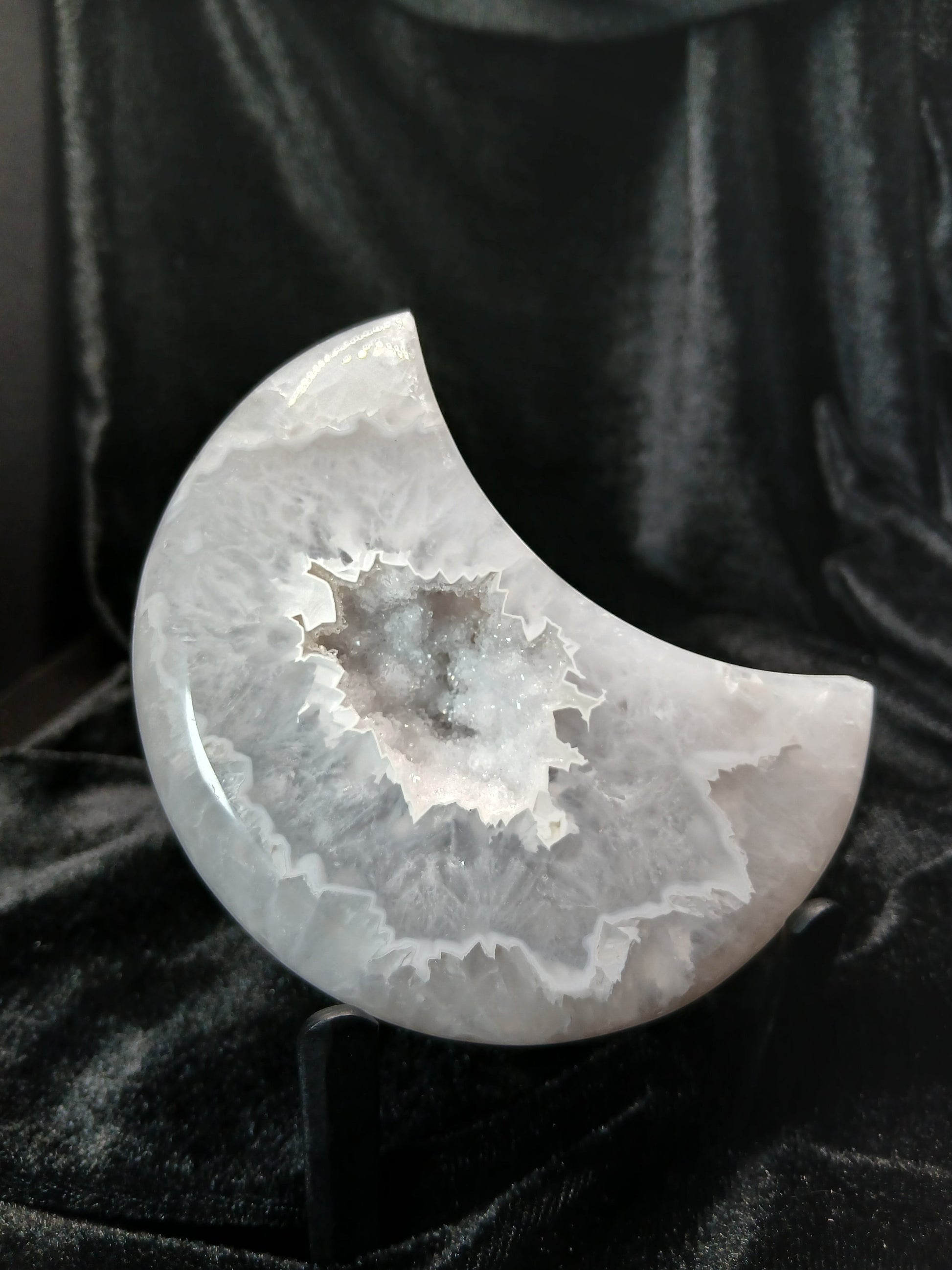Fordite Haus: Crystals & Fordite agate moon carving with a central druzy crystal cluster on a black display stand.