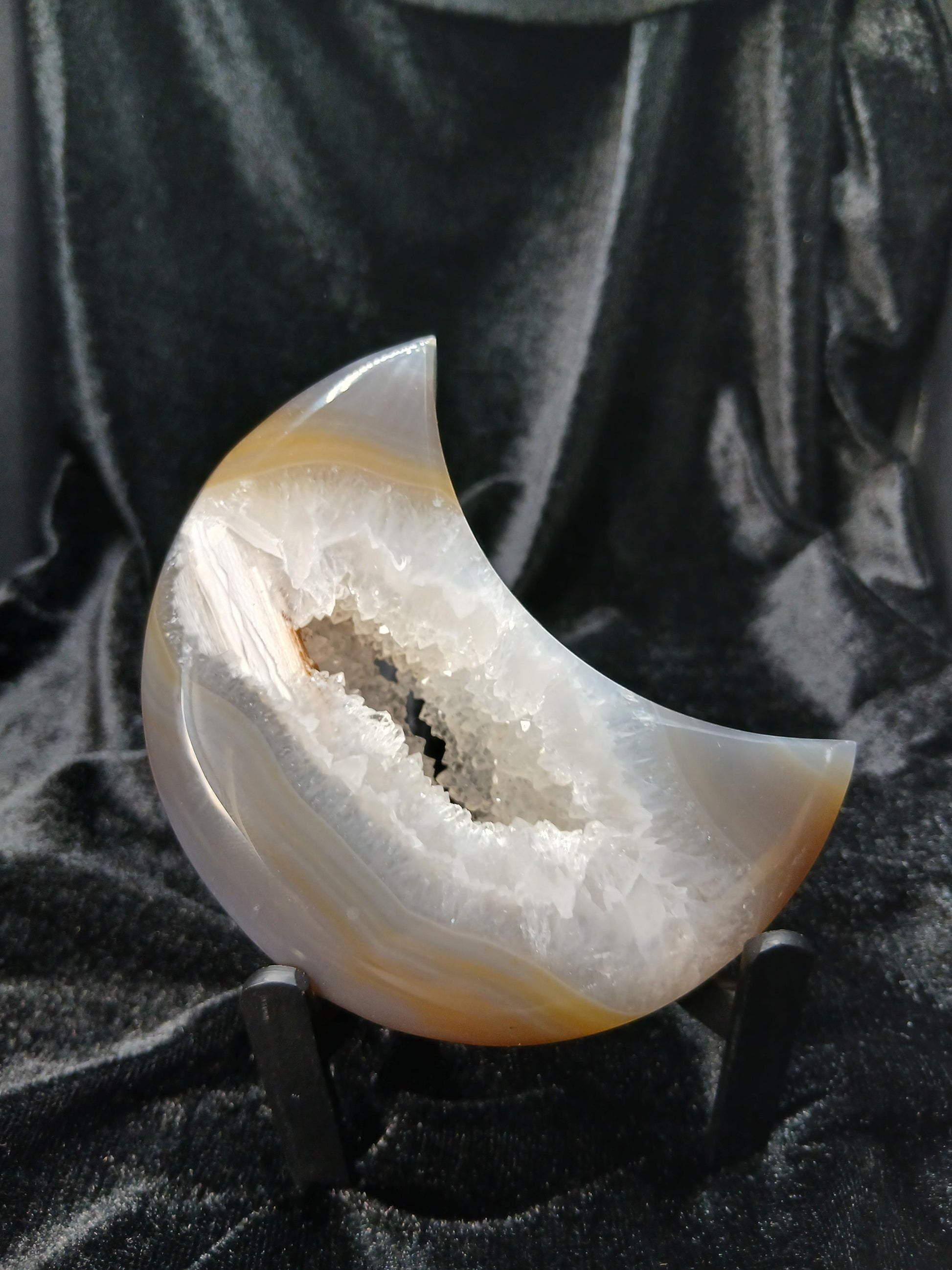 Fordite Haus: Crystals & Fordite agate moon carving with a sparkling crystal geode center on a black display stand.