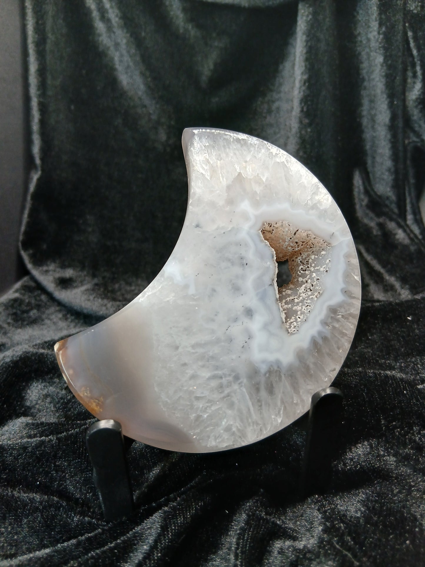 Fordite Haus: Crystals & Fordite agate moon carving with a central druzy geode cavity on a black display stand.
