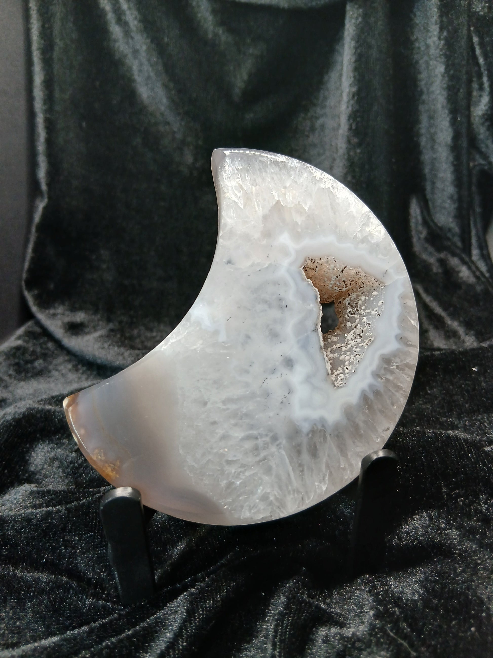 Fordite Haus: Crystals & Fordite agate moon carving with a central druzy geode cavity on a black display stand.
