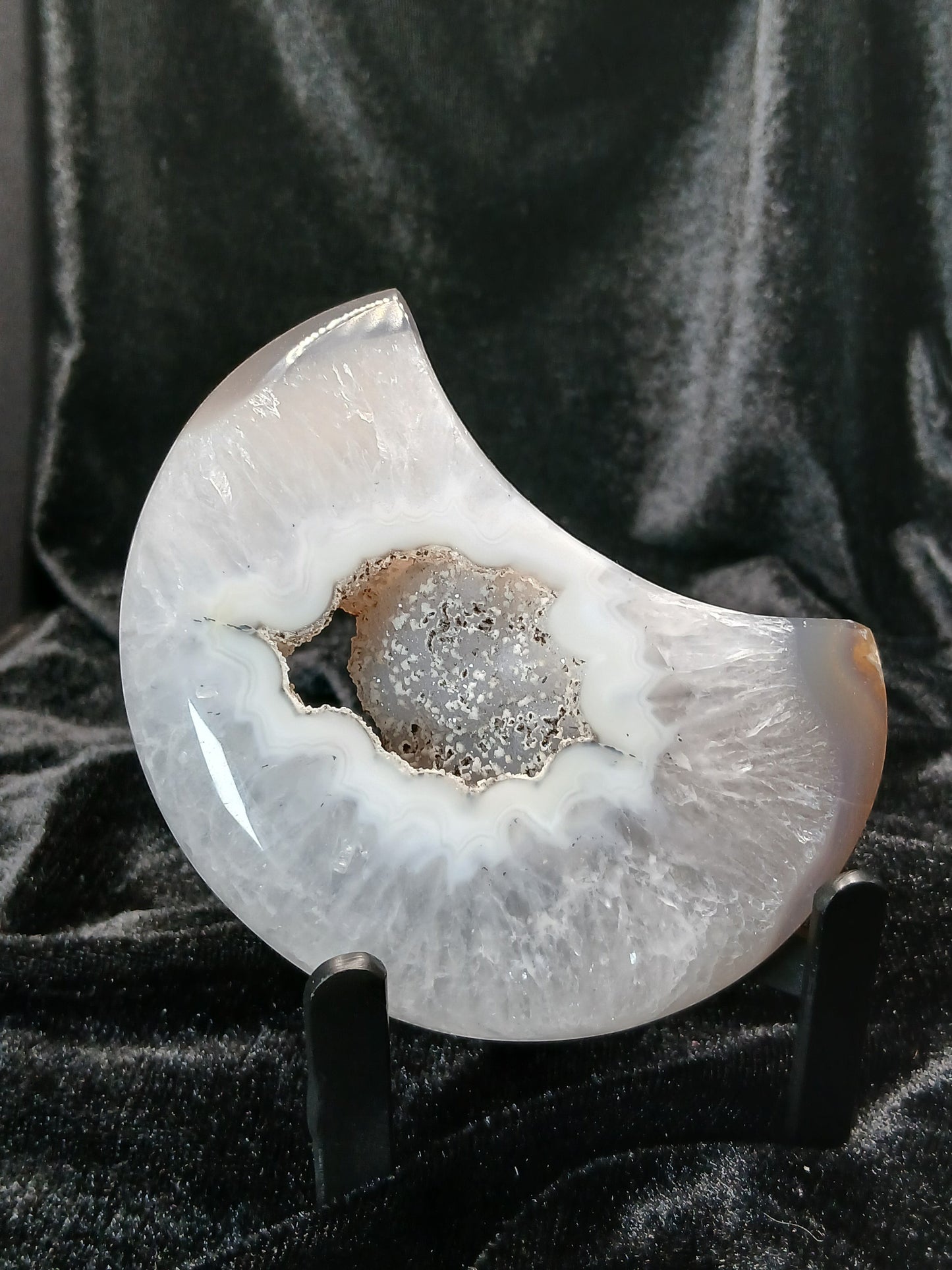 Fordite Haus: Crystals & Fordite agate moon carving with a central druzy geode cavity on a black display stand.
