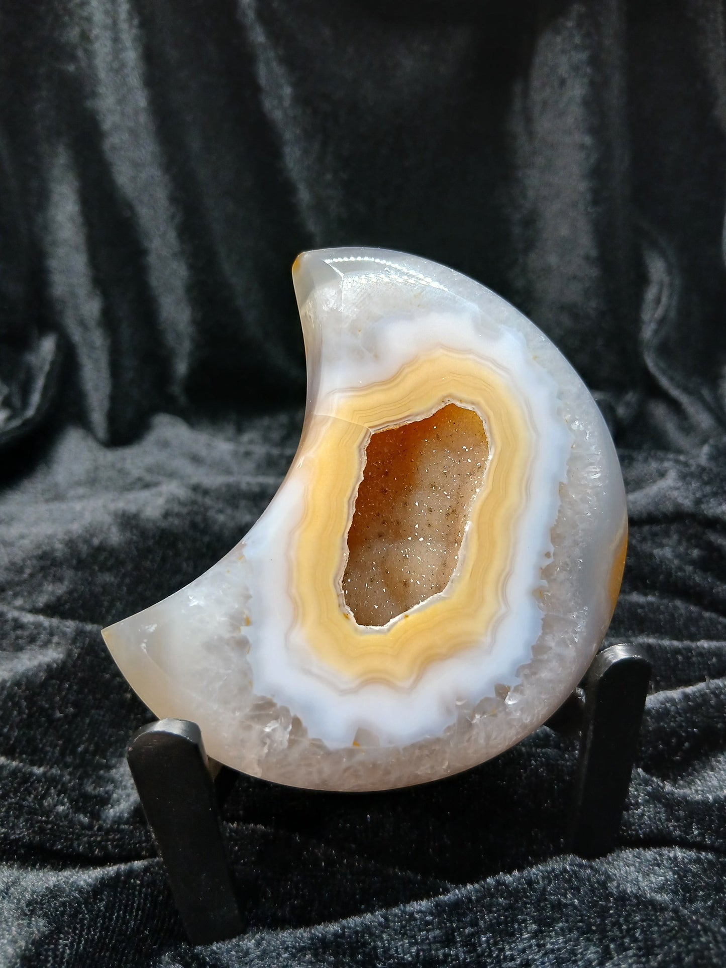 Fordite Haus: Crystals & Fordite peachy agate crescent moon carving with a central druzy geode on a black display stand.