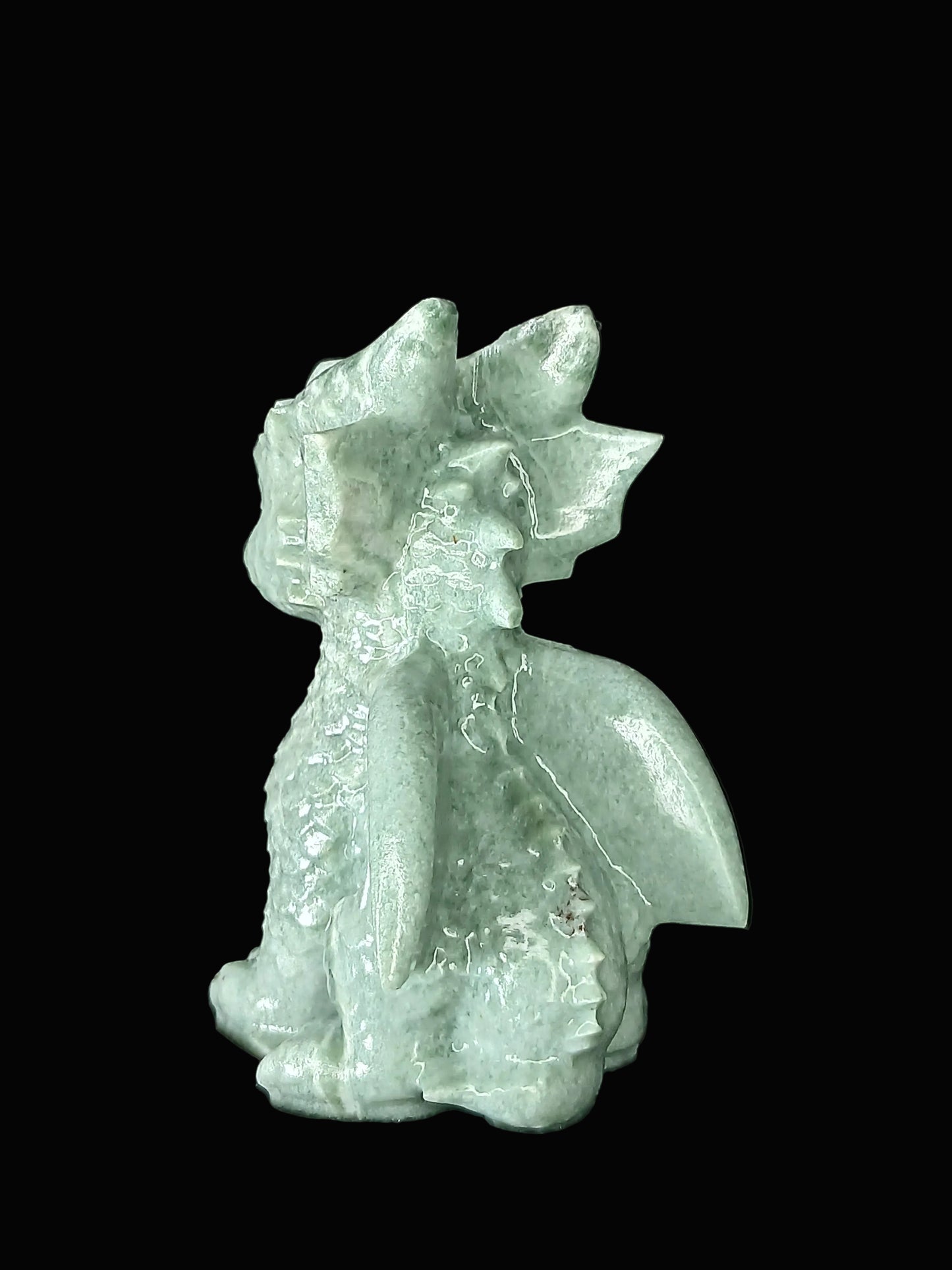 A small carved dragon figurine made of pale green aventurine stone, shown sitting against a dark black fabric background.