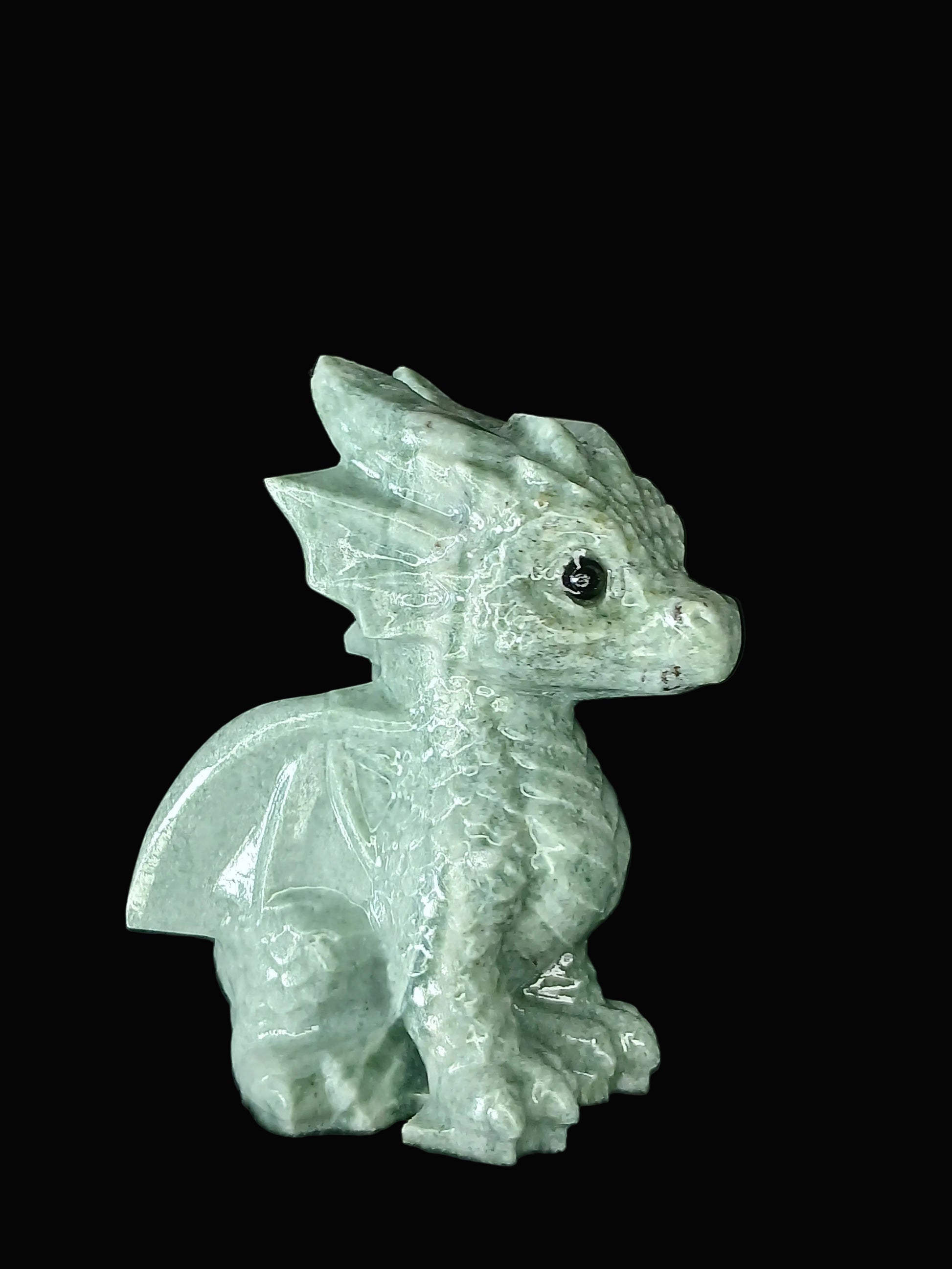 A small carved dragon figurine in pale green aventurine, shown in profile against a dark black fabric background.