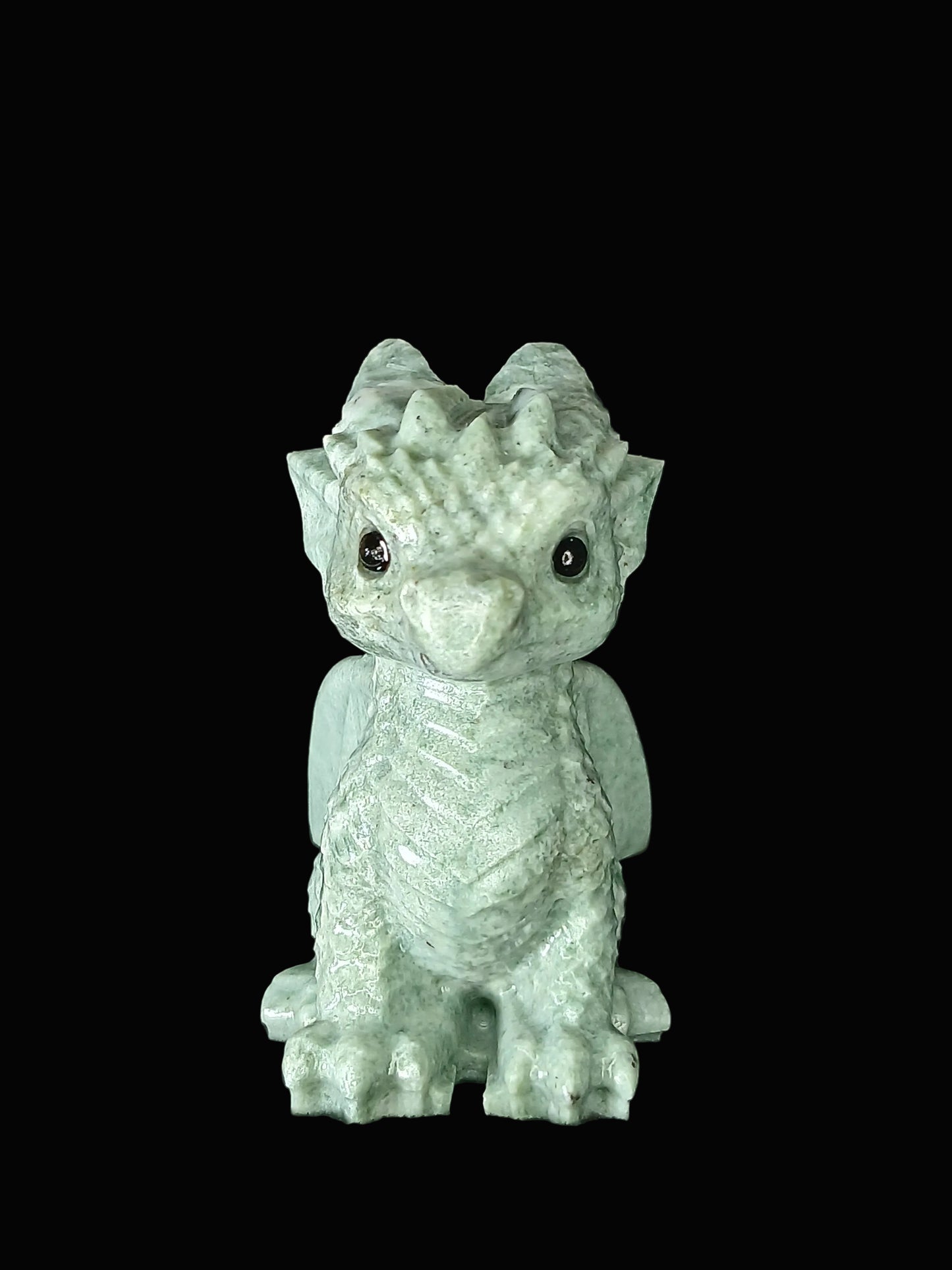 A small pale green dragon figurine with textured scales, large dark eyes, and tiny wings, standing on a dark fabric surface against a black background.
